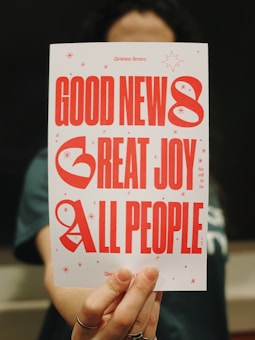 A person is holding a card with the text 'GOOD NEWS, GREAT JOY, ALL PEOPLE' in large red letters. The card is decorated with small red stars and a few lines of smaller text.