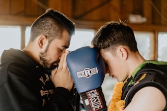Two individuals are facing each other closely, one with closed eyes, in a moment of concentration or meditation. One is holding a blue boxing glove with ‘Ringside’ branding, while the other wears protective gear and a floral garland.