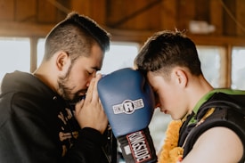 Two individuals are facing each other closely, one with closed eyes, in a moment of concentration or meditation. One is holding a blue boxing glove with ‘Ringside’ branding, while the other wears protective gear and a floral garland.