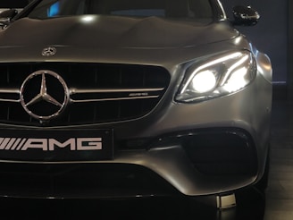 A close-up view of a high-performance luxury car, featuring a prominent Mercedes-Benz emblem on the front grille. The left headlight is glowing brightly, capturing the sleek and elegant design of the vehicle. The AMG badge is visible, indicating the car's sporty capabilities.