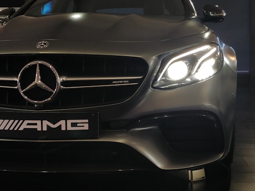 A close-up view of a high-performance luxury car, featuring a prominent Mercedes-Benz emblem on the front grille. The left headlight is glowing brightly, capturing the sleek and elegant design of the vehicle. The AMG badge is visible, indicating the car's sporty capabilities.