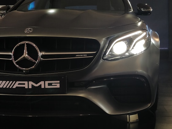 A close-up view of a high-performance luxury car, featuring a prominent Mercedes-Benz emblem on the front grille. The left headlight is glowing brightly, capturing the sleek and elegant design of the vehicle. The AMG badge is visible, indicating the car's sporty capabilities.