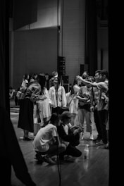 A group of people gather on a stage in what appears to be a backstage or rehearsal setting. Several individuals are standing, while a couple are crouched down, possibly looking at something of interest. The environment is casual, with a mix of casual and long clothing, suggesting a relaxed atmosphere.