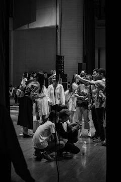 A group of people gather on a stage in what appears to be a backstage or rehearsal setting. Several individuals are standing, while a couple are crouched down, possibly looking at something of interest. The environment is casual, with a mix of casual and long clothing, suggesting a relaxed atmosphere.