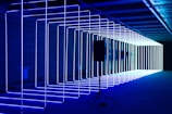 A series of parallel neon light rods create an illuminated tunnel effect in a dimly lit environment. The lights are predominantly blue and white, extending into the distance. A speaker stands on a tripod in the foreground, contrasting with the geometric light structures.