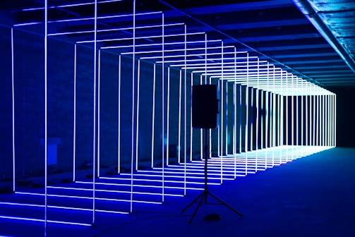 A series of parallel neon light rods create an illuminated tunnel effect in a dimly lit environment. The lights are predominantly blue and white, extending into the distance. A speaker stands on a tripod in the foreground, contrasting with the geometric light structures.