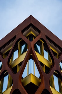 A building with a modern architectural design features a geometric, lattice-like structure made of rust-colored metal framing large, reflective windows. The metal framework creates a striking contrast with the bright yellow accents bordering the windows. The composition creates a symmetrical and dynamic perspective against the backdrop of a clear sky.