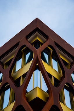 A building with a modern architectural design features a geometric, lattice-like structure made of rust-colored metal framing large, reflective windows. The metal framework creates a striking contrast with the bright yellow accents bordering the windows. The composition creates a symmetrical and dynamic perspective against the backdrop of a clear sky.