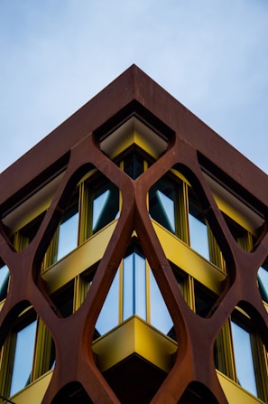 A building with a modern architectural design features a geometric, lattice-like structure made of rust-colored metal framing large, reflective windows. The metal framework creates a striking contrast with the bright yellow accents bordering the windows. The composition creates a symmetrical and dynamic perspective against the backdrop of a clear sky.