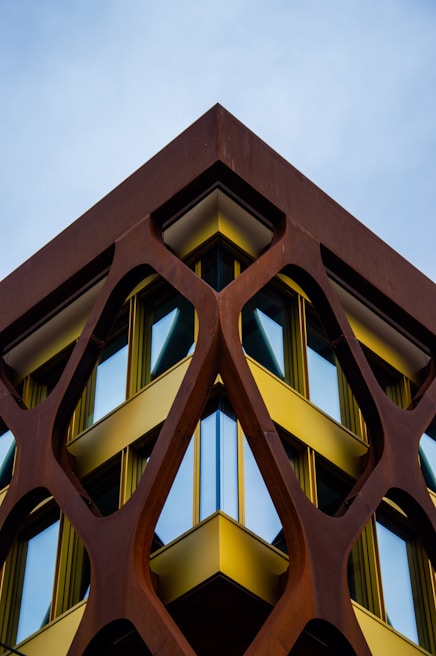 A building with a modern architectural design features a geometric, lattice-like structure made of rust-colored metal framing large, reflective windows. The metal framework creates a striking contrast with the bright yellow accents bordering the windows. The composition creates a symmetrical and dynamic perspective against the backdrop of a clear sky.