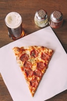 A large slice of pepperoni pizza is placed on a white paper atop a wooden table. Beside the pizza, there is a glass of dark beer with a frothy head. In the background are two shakers, one containing green herbs and the other with red pepper flakes.