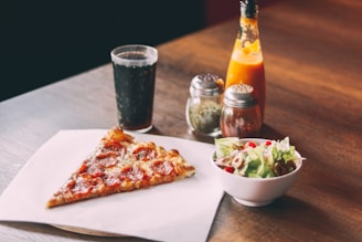 A slice of pepperoni pizza is placed on a piece of parchment paper on a wooden table. Beside it, a small bowl of fresh salad with lettuce, cherry tomatoes, and olives is present. In the background, a glass filled with a dark carbonated drink can be seen next to a bottle of hot sauce and two shakers containing herbs and spices.