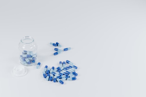A clear glass jar, partially opened with its lid beside it, contains blue and white capsules. Several additional capsules are scattered around the jar on a clean, light background.