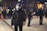 Police officers in tactical gear are standing in an urban public space at night. They are wearing helmets and holding riot shields, suggesting a preparation for or response to a protest or similar situation. The background is blurred, with city lights illuminating the scene and other officers visible.