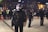 Police officers in tactical gear are standing in an urban public space at night. They are wearing helmets and holding riot shields, suggesting a preparation for or response to a protest or similar situation. The background is blurred, with city lights illuminating the scene and other officers visible.