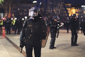 Police officers in tactical gear are standing in an urban public space at night. They are wearing helmets and holding riot shields, suggesting a preparation for or response to a protest or similar situation. The background is blurred, with city lights illuminating the scene and other officers visible.