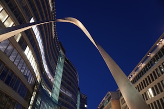 An architectural scene featuring tall office buildings with illuminated windows. Curved structures and modern glass and metal elements dominate the composition, set against a clear evening sky.