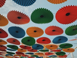 Multiple colorful parasols are suspended in the air, arranged in a grid pattern against a clear blue sky. The parasols consist of different circular pleated designs in red, blue, green, yellow, and orange, creating a vibrant canopy.