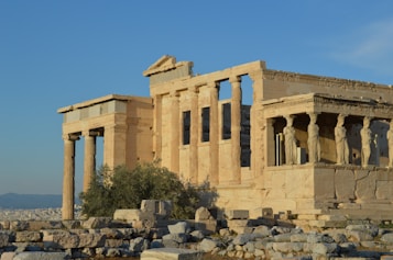 Ancient temple ruins with classical Greek architectural style, featuring tall columns and statues integrated into the structure. The stone construction reflects a light beige color under the sunlight. The background reveals a landscape view with a city and distant hills.