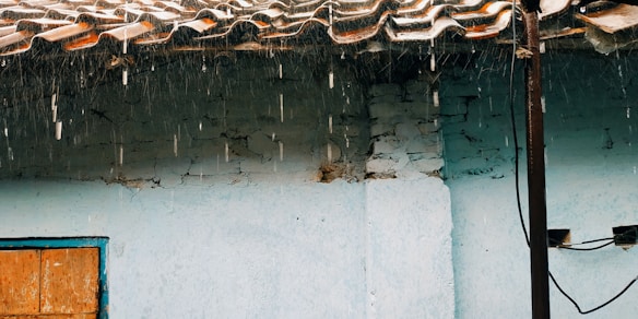 A rustic structure with a tiled roof and a light blue wall is shown with rainwater dripping from the roof. The wall has visible cracks and peeling paint. A wooden window frame with some cables is noticeable on the side.