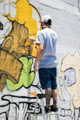 A person stands on a ladder, applying yellow spray paint to a wall covered in graffiti art. The artwork features bold outlines and abstract shapes with a mix of green, yellow, and black colors.