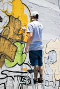 A person stands on a ladder, applying yellow spray paint to a wall covered in graffiti art. The artwork features bold outlines and abstract shapes with a mix of green, yellow, and black colors.
