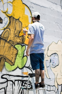 A person stands on a ladder, applying yellow spray paint to a wall covered in graffiti art. The artwork features bold outlines and abstract shapes with a mix of green, yellow, and black colors.