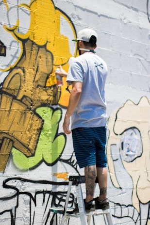 A person stands on a ladder, applying yellow spray paint to a wall covered in graffiti art. The artwork features bold outlines and abstract shapes with a mix of green, yellow, and black colors.