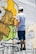 A person stands on a ladder, applying yellow spray paint to a wall covered in graffiti art. The artwork features bold outlines and abstract shapes with a mix of green, yellow, and black colors.