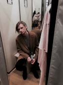 A person is sitting in a fitting room wearing a brown coat with a price tag attached to it. The individual appears to have a contemplative expression. The room is small, with mirrors reflecting the image, and there are hanging clothes visible.
