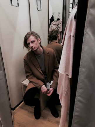 A person is sitting in a fitting room wearing a brown coat with a price tag attached to it. The individual appears to have a contemplative expression. The room is small, with mirrors reflecting the image, and there are hanging clothes visible.