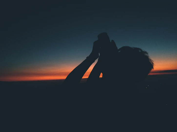 A trekker silhouetted against a sunset, peering through high-powered binoculars over jagged mountain peaks.