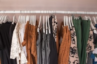 A clothing rack displaying various garments on white hangers. The clothes include dresses, shirts, and pants in a mix of patterns and solid colors such as brown, black, white, green, and gray. Floral and abstract designs are visible among the clothes, creating a visually appealing assortment.