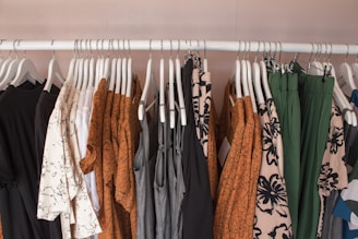 A clothing rack displaying various garments on white hangers. The clothes include dresses, shirts, and pants in a mix of patterns and solid colors such as brown, black, white, green, and gray. Floral and abstract designs are visible among the clothes, creating a visually appealing assortment.