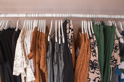A clothing rack displaying various garments on white hangers. The clothes include dresses, shirts, and pants in a mix of patterns and solid colors such as brown, black, white, green, and gray. Floral and abstract designs are visible among the clothes, creating a visually appealing assortment.