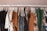 A clothing rack displaying various garments on white hangers. The clothes include dresses, shirts, and pants in a mix of patterns and solid colors such as brown, black, white, green, and gray. Floral and abstract designs are visible among the clothes, creating a visually appealing assortment.