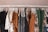 A clothing rack displaying various garments on white hangers. The clothes include dresses, shirts, and pants in a mix of patterns and solid colors such as brown, black, white, green, and gray. Floral and abstract designs are visible among the clothes, creating a visually appealing assortment.