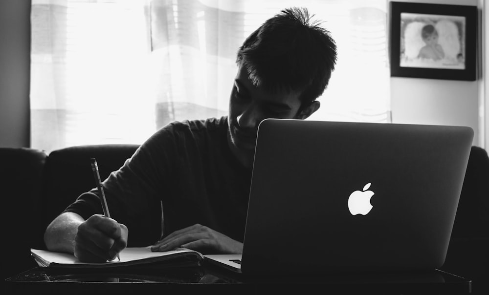 grayscale photography of man writing in front of MacBook