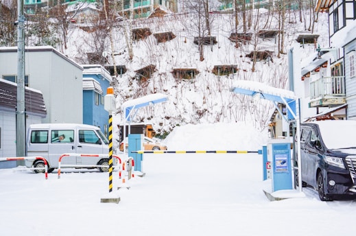 A snowy parking lot with entry barriers. Two vehicles, a silver van on the left and a black van on the right, are parked. The area is surrounded by buildings and a snow-covered hill with sparse trees and structures. There are control barriers at the entrance and caution poles painted yellow and black.