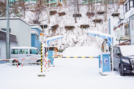 A snowy parking lot with entry barriers. Two vehicles, a silver van on the left and a black van on the right, are parked. The area is surrounded by buildings and a snow-covered hill with sparse trees and structures. There are control barriers at the entrance and caution poles painted yellow and black.