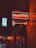 A neon sign reading 'Newsstand' is displayed with bright orange and blue colors. The sign is mounted on a wall in an indoor setting with reflections of neon lights on the shiny surface. The surrounding area appears dimly lit, enhancing the sign's glow.