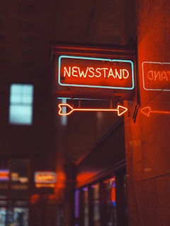 A neon sign reading 'Newsstand' is displayed with bright orange and blue colors. The sign is mounted on a wall in an indoor setting with reflections of neon lights on the shiny surface. The surrounding area appears dimly lit, enhancing the sign's glow.