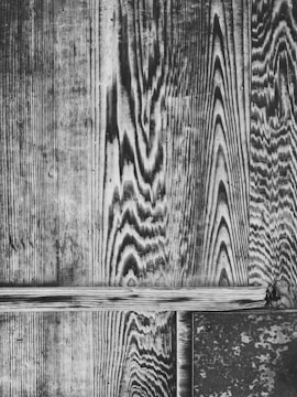 The image displays a detailed view of a vintage wooden surface with distinct grain patterns. The wood appears aged, showcasing a mix of linear and wavy textures that create an intricate design. At the bottom, a metal piece is slightly visible, contrasting with the wood.