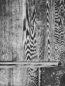The image displays a detailed view of a vintage wooden surface with distinct grain patterns. The wood appears aged, showcasing a mix of linear and wavy textures that create an intricate design. At the bottom, a metal piece is slightly visible, contrasting with the wood.