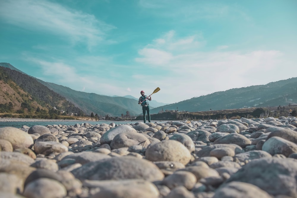 man holding oar standing on stones near body of water during daytime