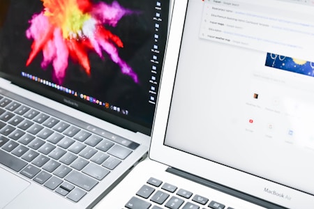 Two laptops are positioned closely side by side. The left laptop displays a vibrant, colorful abstract image, while the right laptop shows a web browser with a search engine page open. The keyboards and part of the screens are visible.