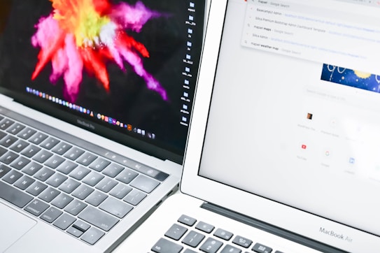 Two laptops are positioned closely side by side. The left laptop displays a vibrant, colorful abstract image, while the right laptop shows a web browser with a search engine page open. The keyboards and part of the screens are visible.