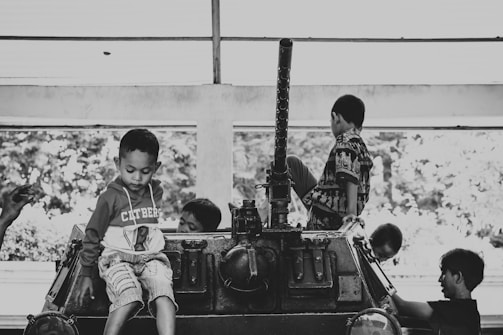 Children are sitting and playing on top of a stationary military vehicle. The scene is captured in black and white, highlighting the contrast between the children's playful demeanor and the serious nature of the vehicle. The setting appears to be indoors with a large window in the background.