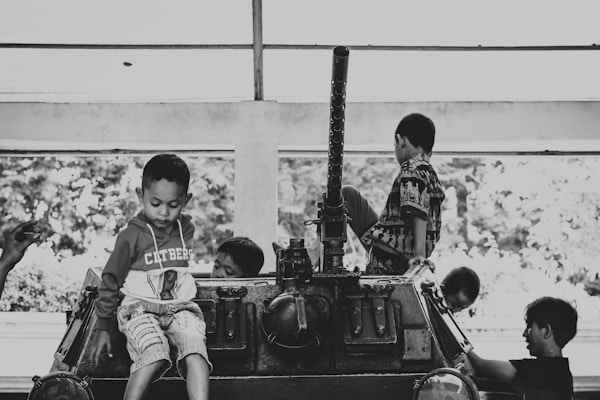 Children are sitting and playing on top of a stationary military vehicle. The scene is captured in black and white, highlighting the contrast between the children's playful demeanor and the serious nature of the vehicle. The setting appears to be indoors with a large window in the background.