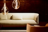 A cozy and modern living space featuring a light-colored sofa with a matching pillow in front of a dark brick wall. Two large, stylish pendant lights with visible filaments hang from the ceiling, casting a warm glow. An armchair in complementary tones is situated nearby.